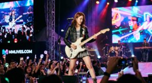 Nữ nghệ sĩ trẻ mặc áo khoác da đen đang chơi đàn guitar điện và hát trên sân khấu lớn tại một buổi concert. Màn hình LED phía sau hiển thị logo 'MMLIVES.COM'. Khán giả trong foreground giơ cao lightstick và điện thoại.